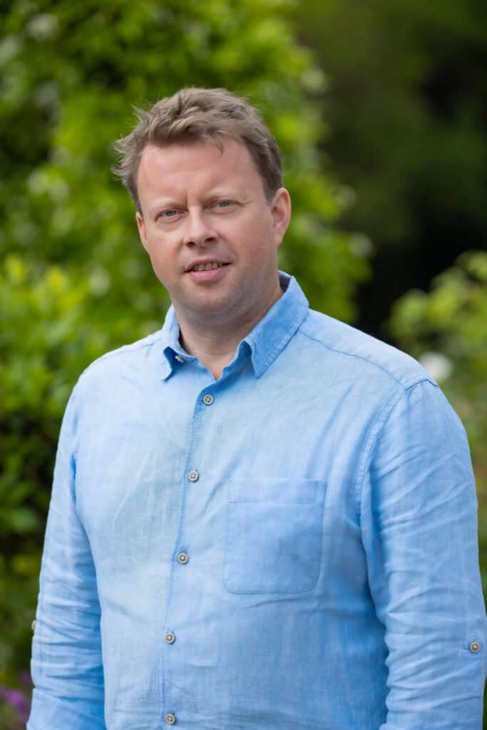 A man with light brown hair and a light blue shirt looks directly at the viewer, with a blurred green background.