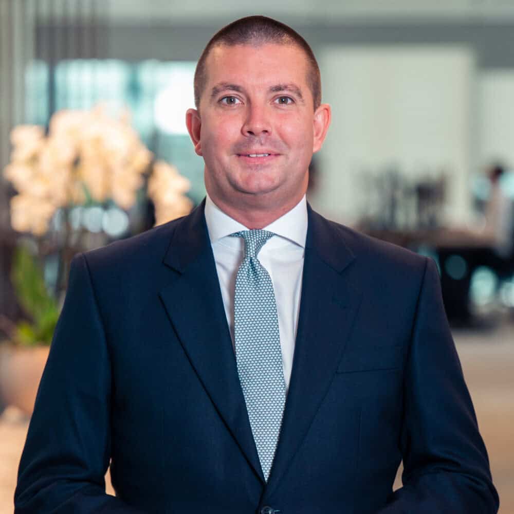 A man in a dark suit and light blue tie smiles at the camera, standing in a modern office.