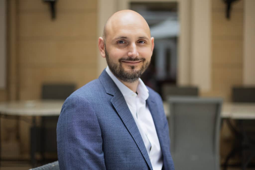 Headshot of a bald man with a beard, smiling, wearing a blue blazer and white shirt.