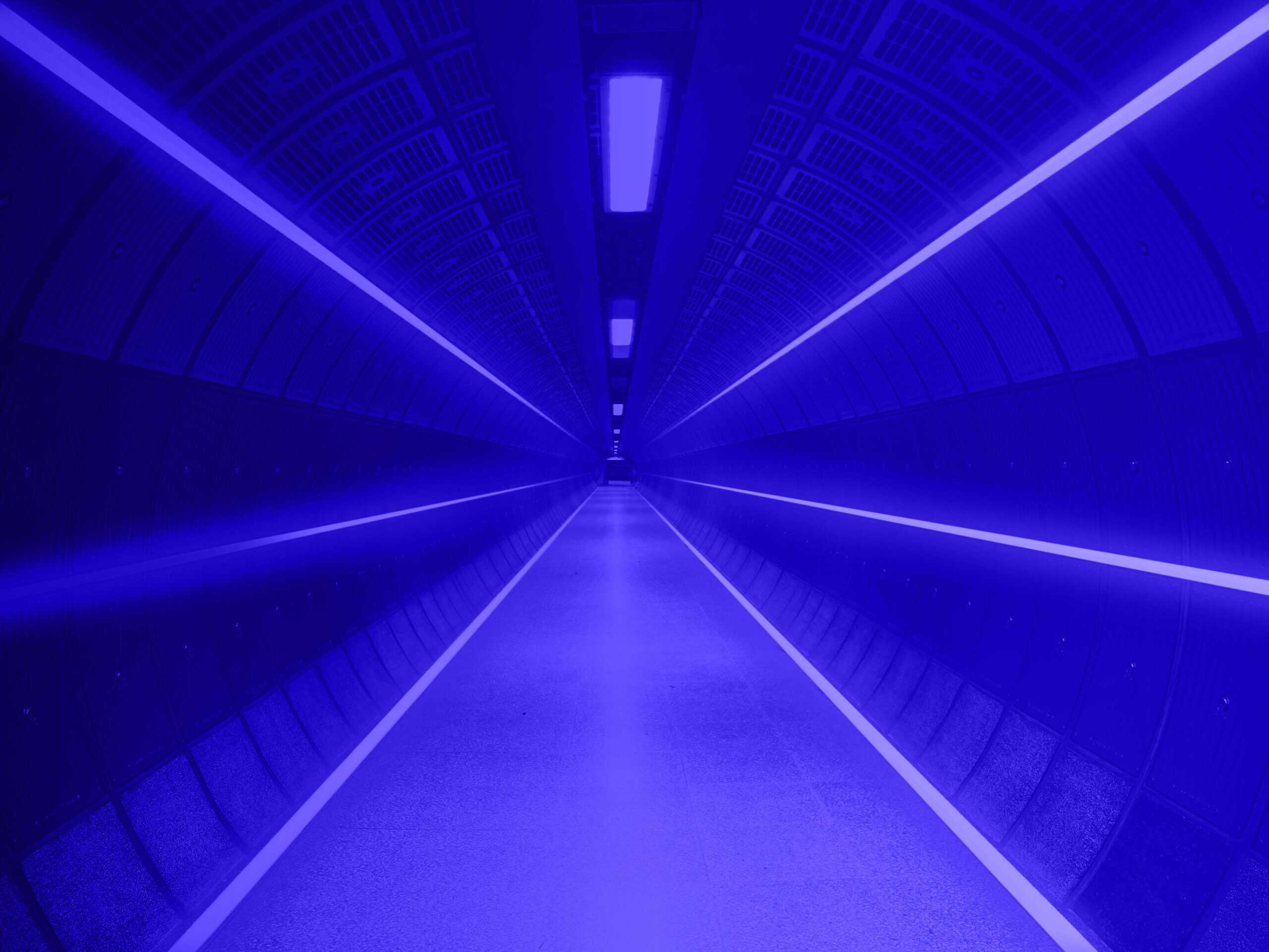 A long, empty tunnel bathed in vibrant blue light, with linear lights on the walls and ceiling, and two white lines on the floor, creating a futuristic perspective.