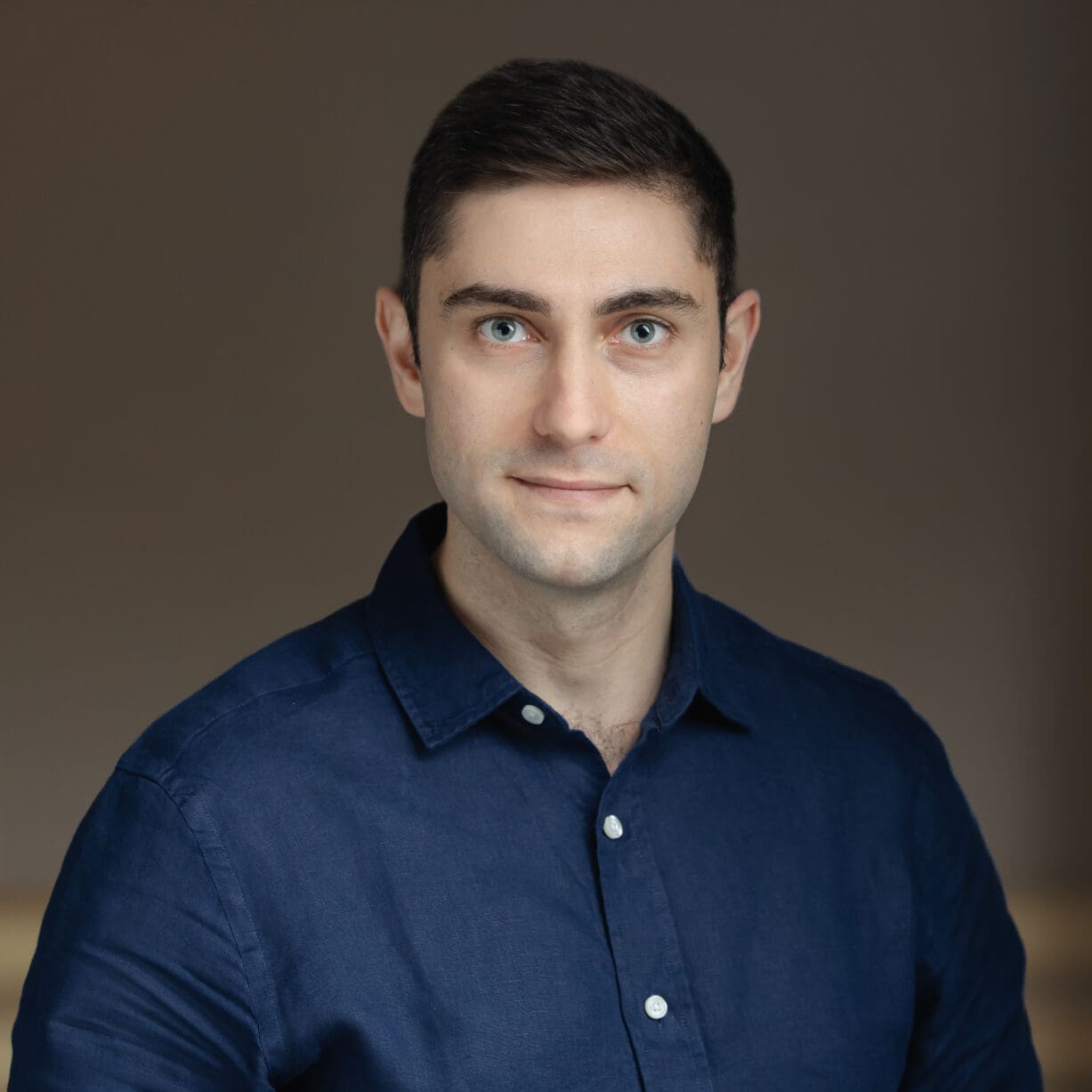 Headshot of a man with dark hair and blue eyes, wearing a dark blue shirt, against a blurred background with a warm light.