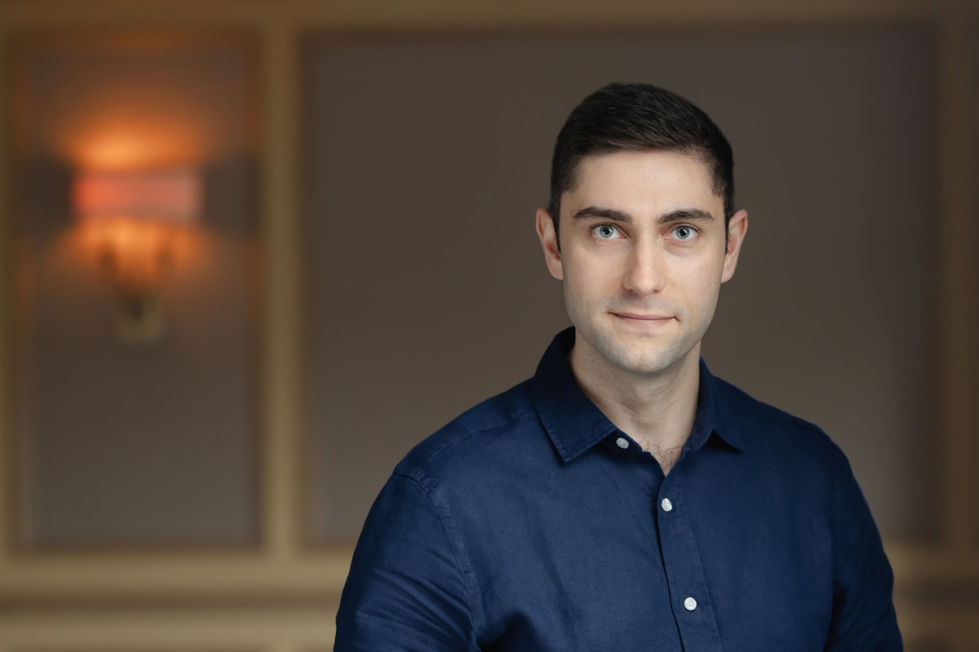 Headshot of a man with dark hair and blue eyes, wearing a dark blue shirt, against a blurred background with a warm light.