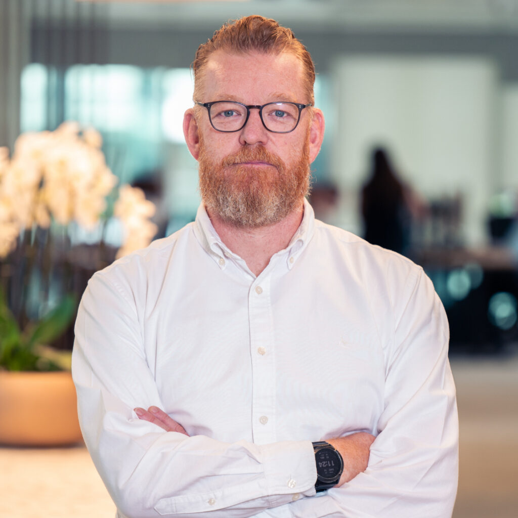 A man with red hair, a red beard, and glasses, wearing a white shirt, stands with his arms crossed in an office.