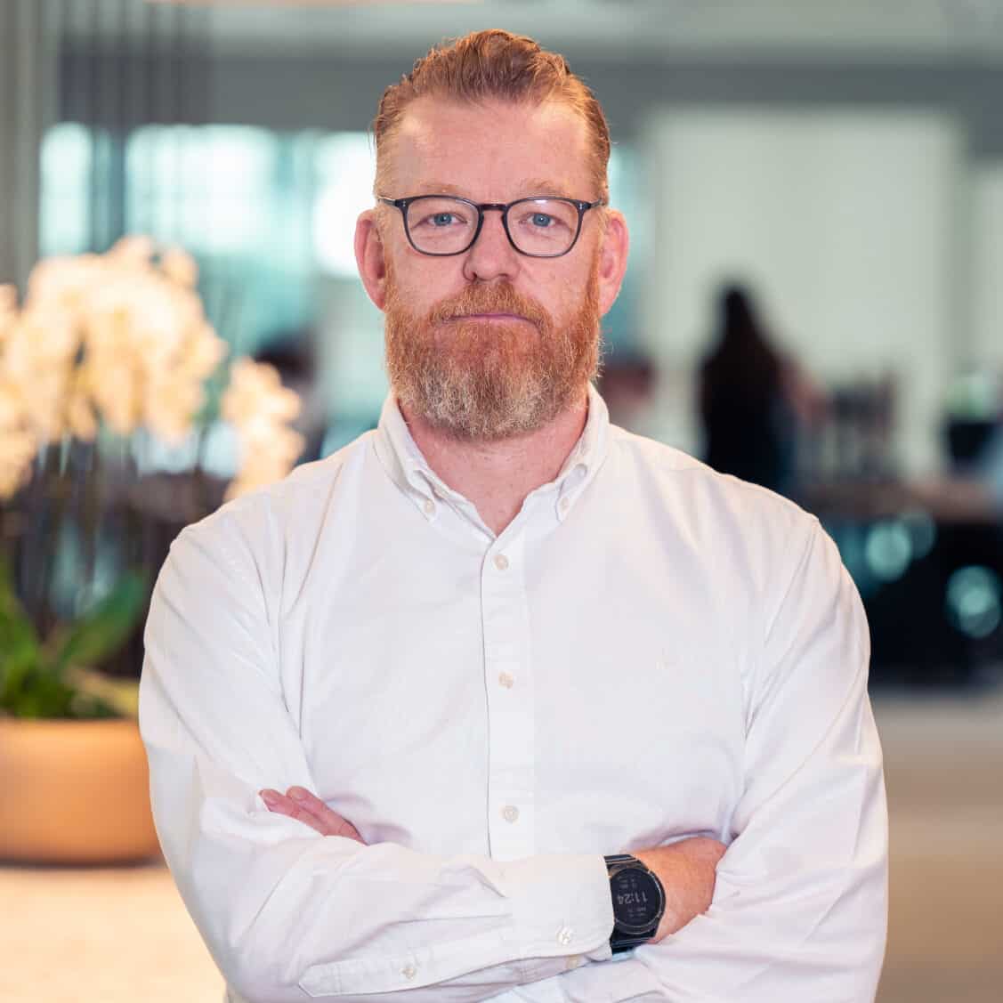 A man with red hair, a red beard, and glasses, wearing a white shirt, stands with his arms crossed in an office.