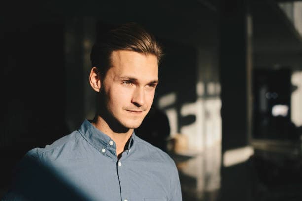 A young man with light brown hair and a blue shirt, illuminated by sunlight on his right side, looks thoughtfully to the right.