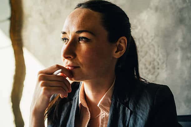 Close-up of a woman with dark hair, looking thoughtfully to her left, holding a white pen to her chin. She wears a dark blazer and pink shirt.