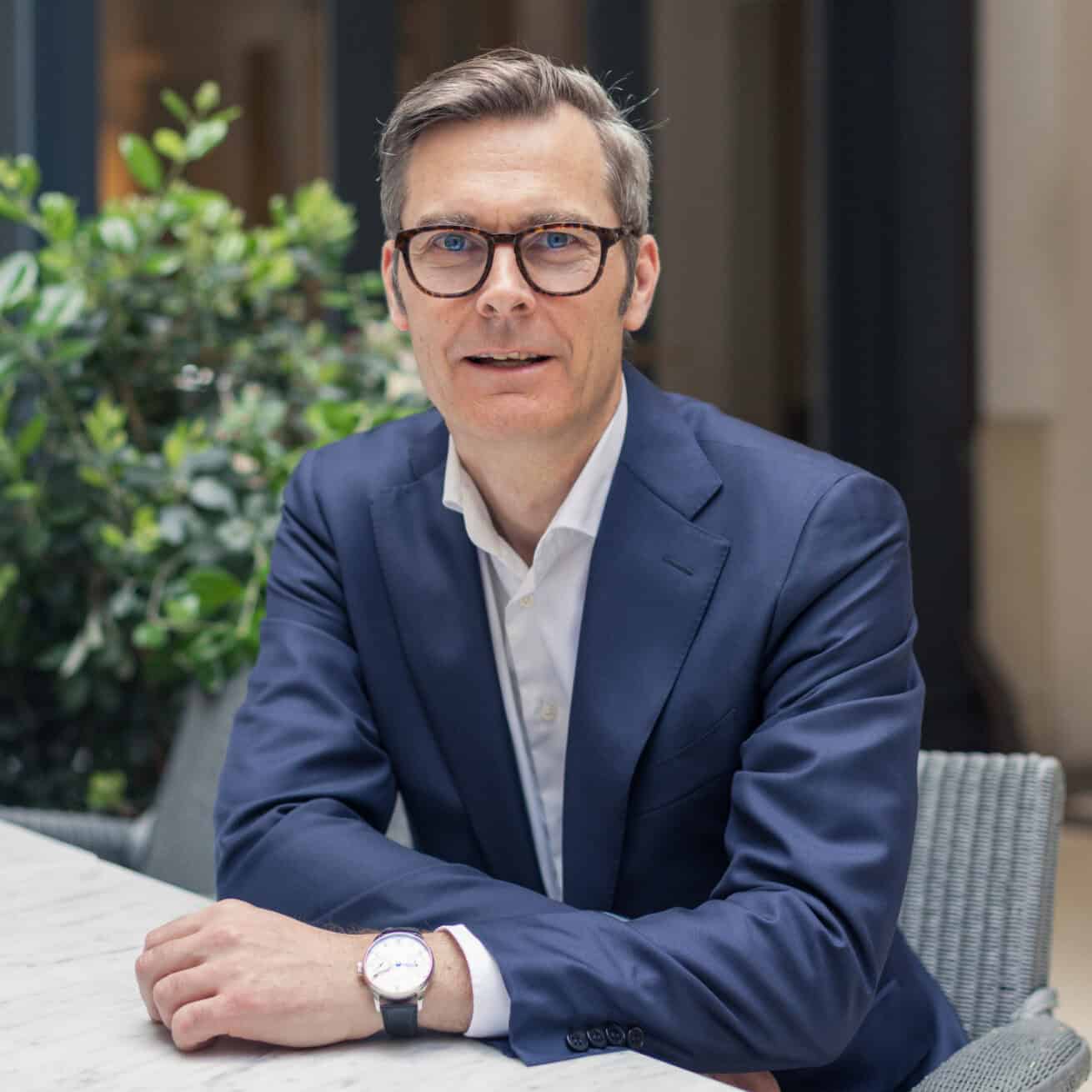 A man in a navy suit and glasses sits at a marble table, looking at the camera.