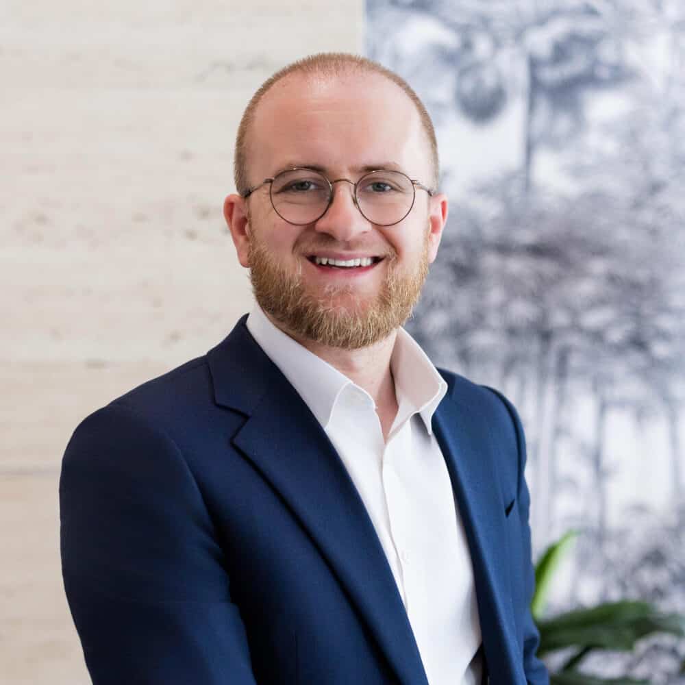 A smiling man with a beard and glasses in a blue suit jacket and white shirt, standing in front of a textured wall and patterned wallpaper.