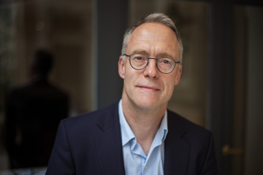Headshot of a man with graying hair and glasses, wearing a blue shirt and dark suit jacket, looking at the camera.