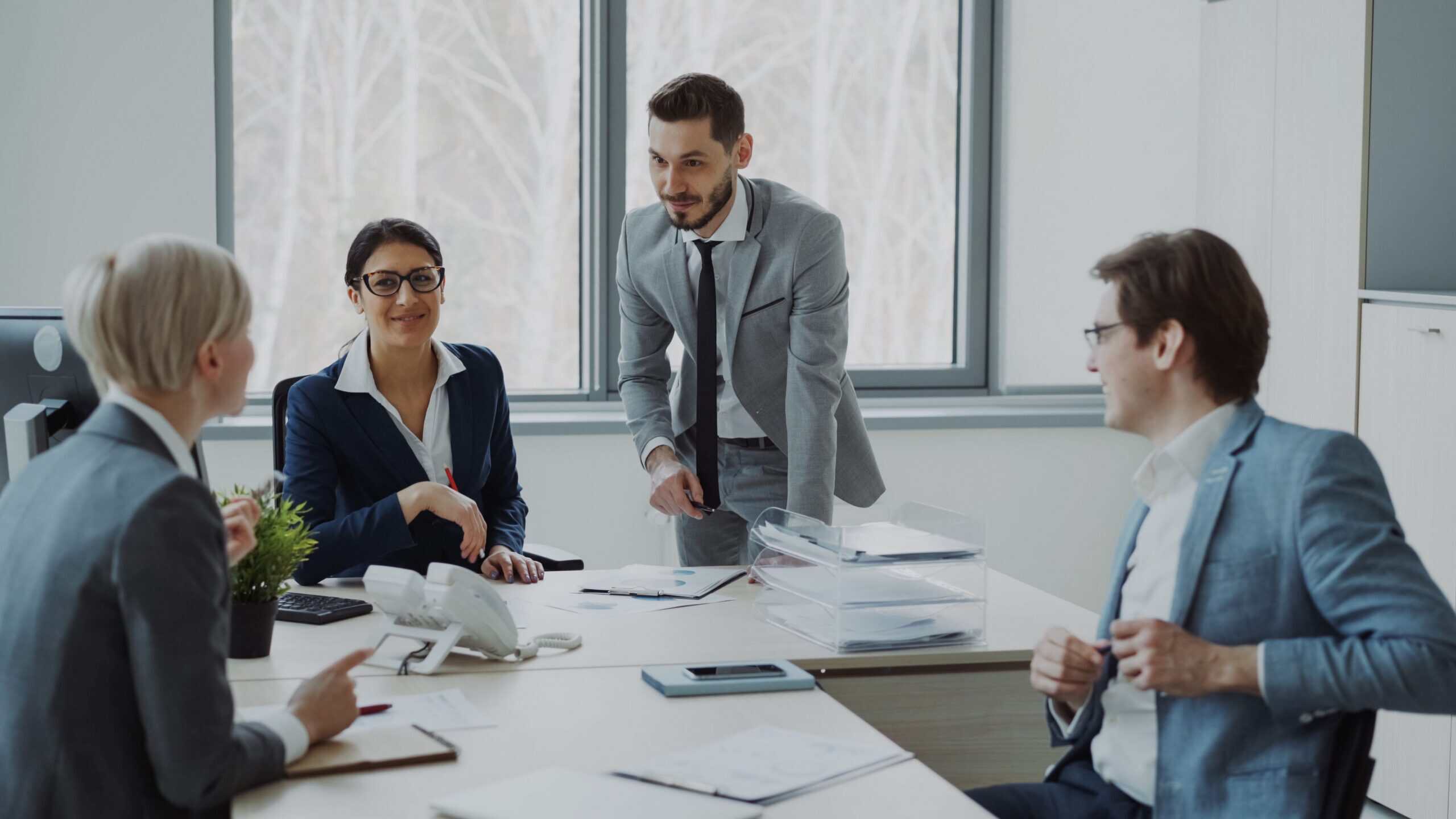 Four business professionals, two men and two women, in suits, are seated and standing around a conference table in an office, engaged in a meeting. Papers and office supplies are on the table.