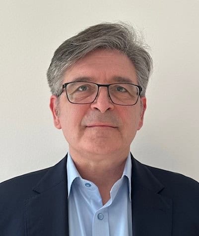 Headshot of a man with graying hair and glasses, wearing a blue shirt and dark blazer.