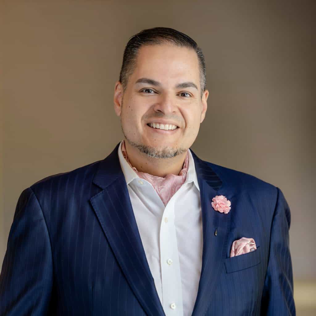 A smiling man in a navy pinstripe suit, white shirt, pink paisley cravat, and matching lapel pin and pocket square.