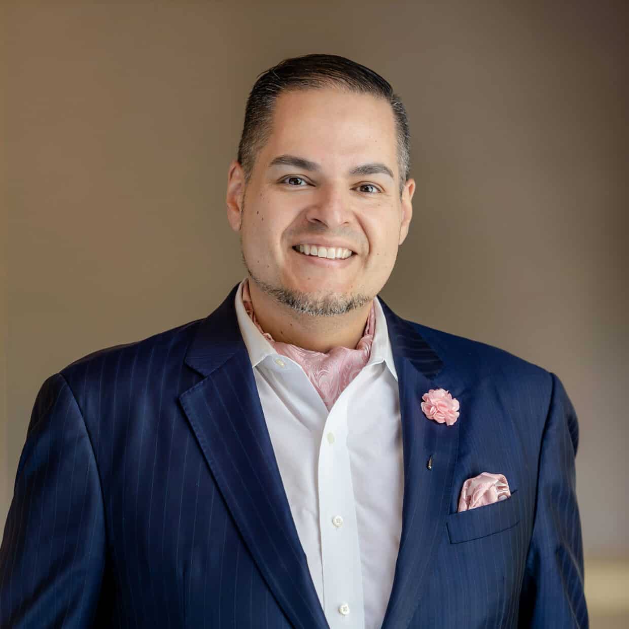 A smiling man in a navy pinstripe suit, white shirt, pink paisley cravat, and matching lapel pin and pocket square.