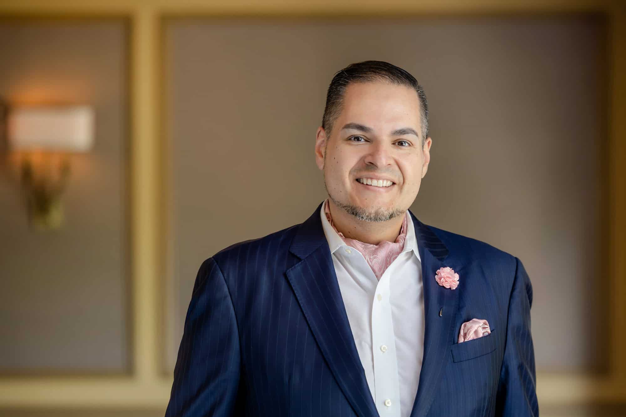 A smiling man in a navy pinstripe suit, white shirt, pink paisley cravat, and matching lapel pin and pocket square.