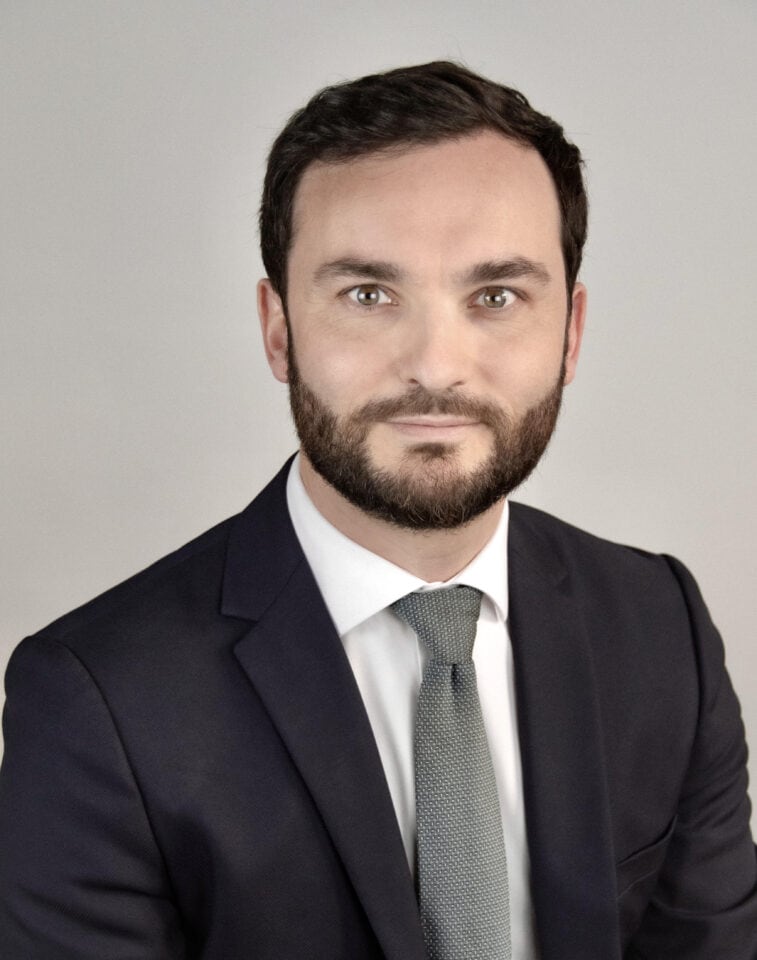 Headshot of a man with dark hair and beard, wearing a navy suit, white shirt, and grey-green tie, against a light grey background.