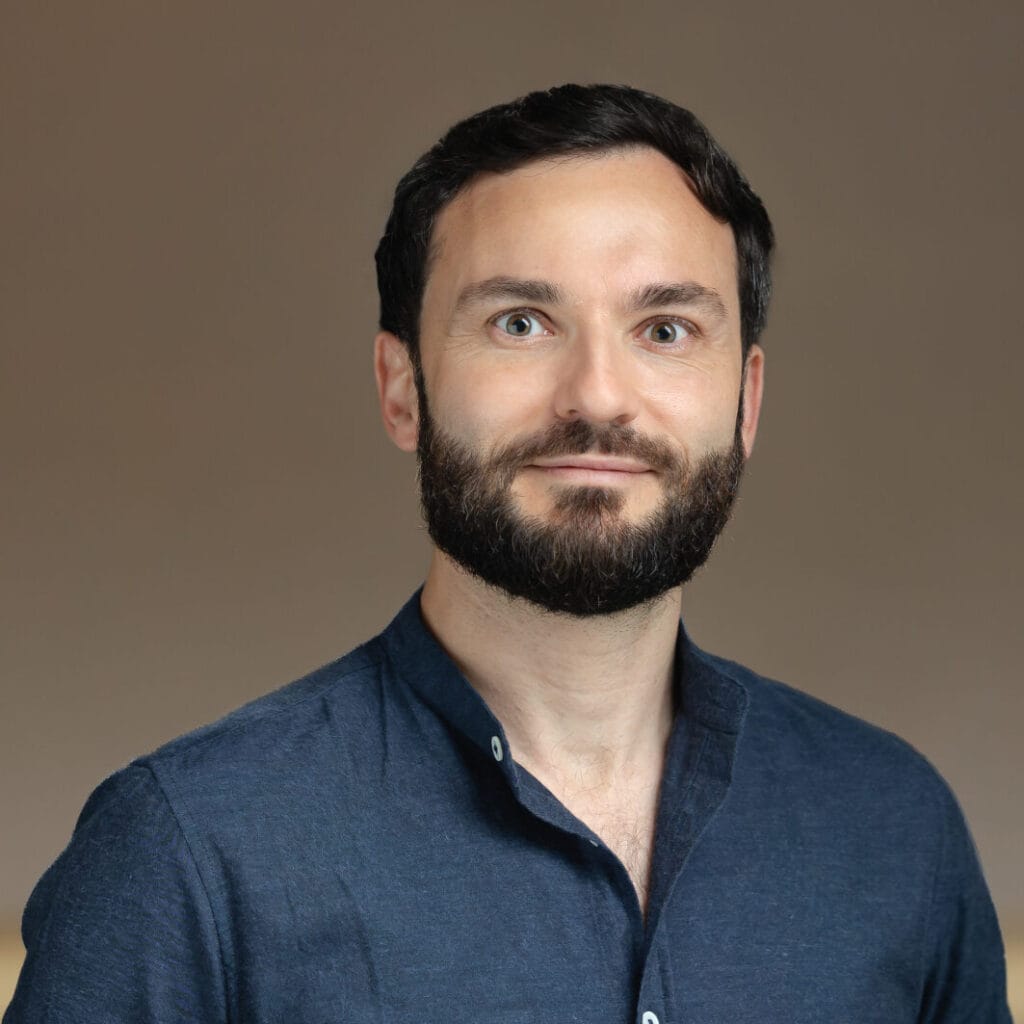 Headshot of a smiling man with dark hair and beard, wearing a dark blue shirt, against a blurred background.
