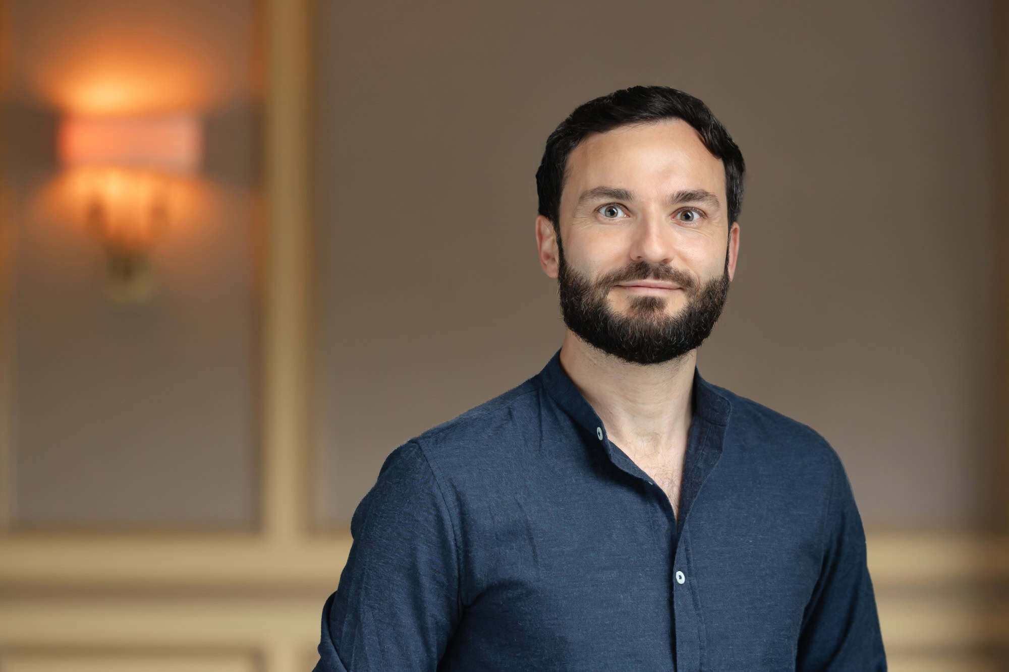 Headshot of a smiling man with dark hair and beard, wearing a dark blue shirt, against a blurred background.