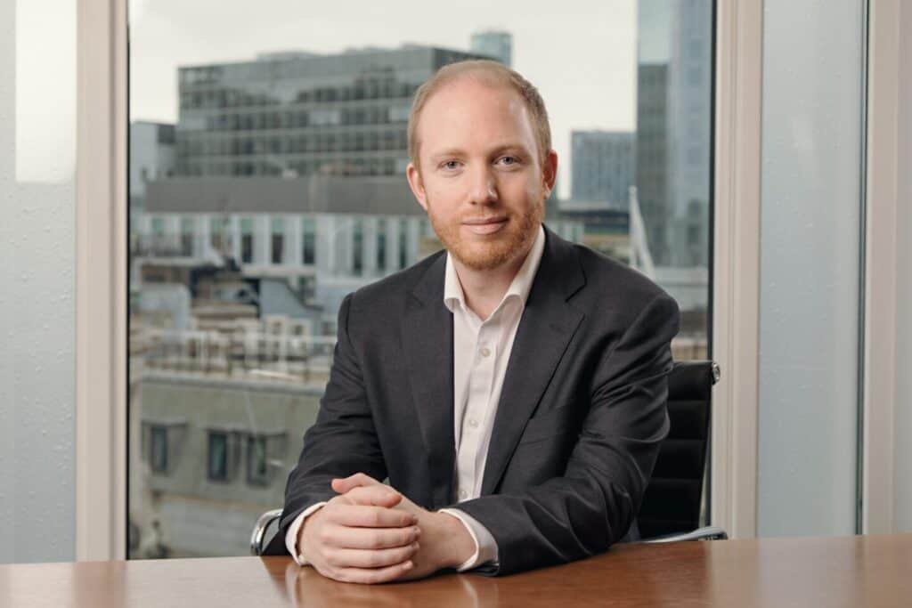 A man with light red hair and a beard, wearing a dark suit, sits at a wooden desk with his hands clasped. A city skyline is visible through a large window behind him.