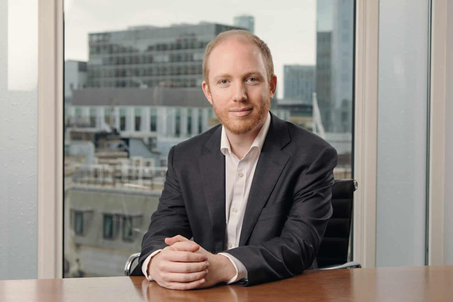 A man with light red hair and a beard, wearing a dark suit, sits at a wooden desk with his hands clasped. A city skyline is visible through a large window behind him.