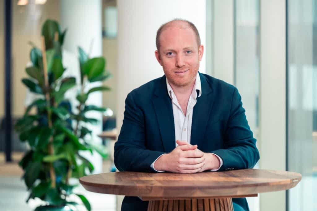 A man with red hair and beard, wearing a dark suit jacket and white shirt, sits at a wooden table with clasped hands, smiling at the camera. A green plant and blurred office background are visible.