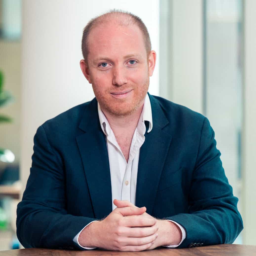 A man with red hair and beard, wearing a dark suit jacket and white shirt, sits at a wooden table with clasped hands, smiling at the camera. A green plant and blurred office background are visible.