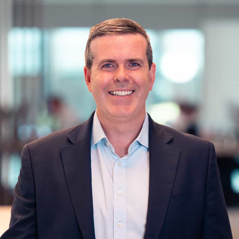 A smiling man in a dark suit jacket and light blue shirt, standing in a blurred modern office background with a white orchid plant visible on the left.