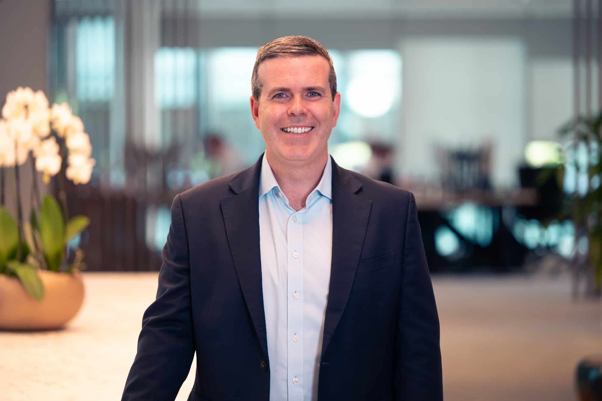 A smiling man in a dark suit jacket and light blue shirt, standing in a blurred modern office background with a white orchid plant visible on the left.