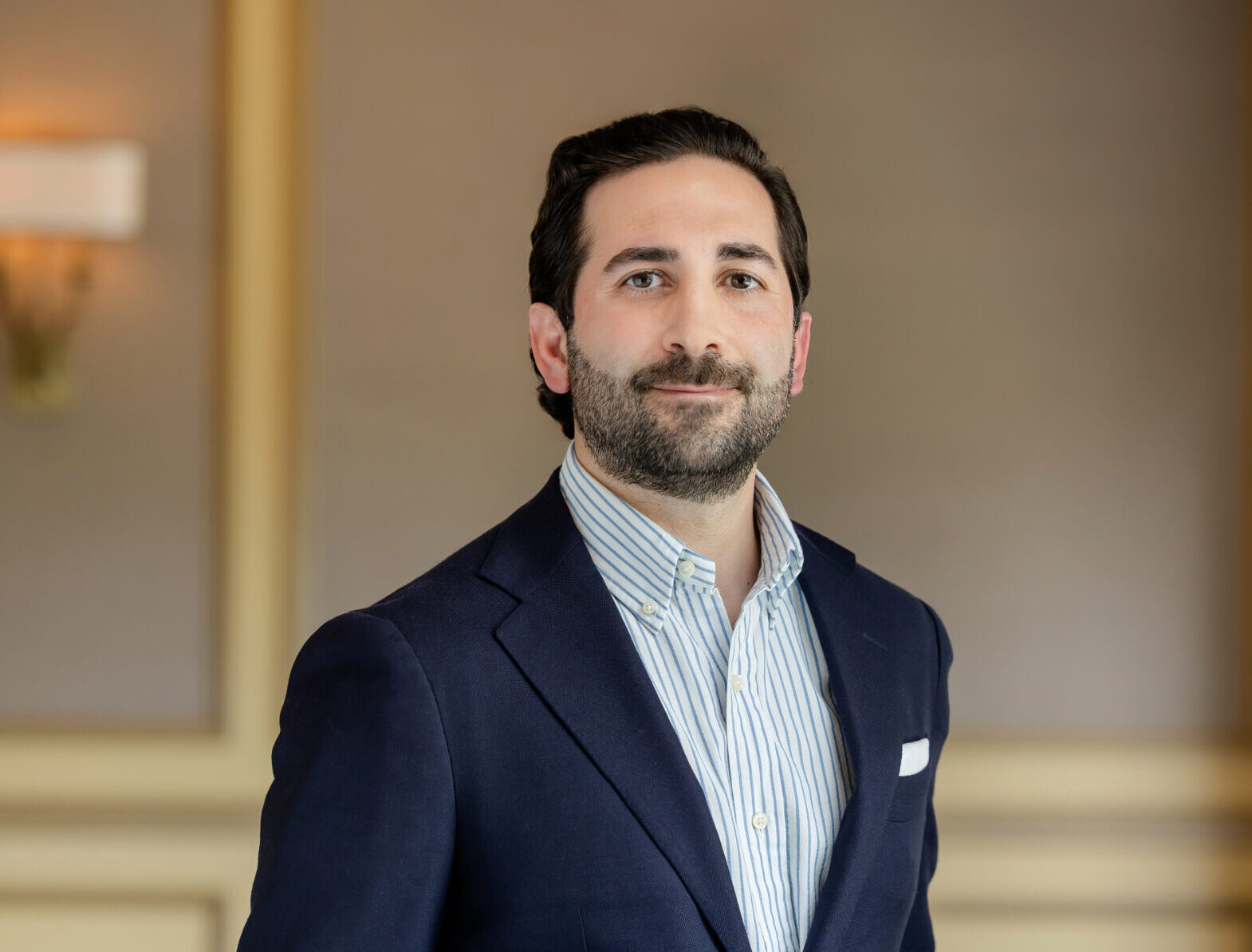A man with dark hair and a beard, wearing a navy blazer and striped shirt, smiles at the camera.