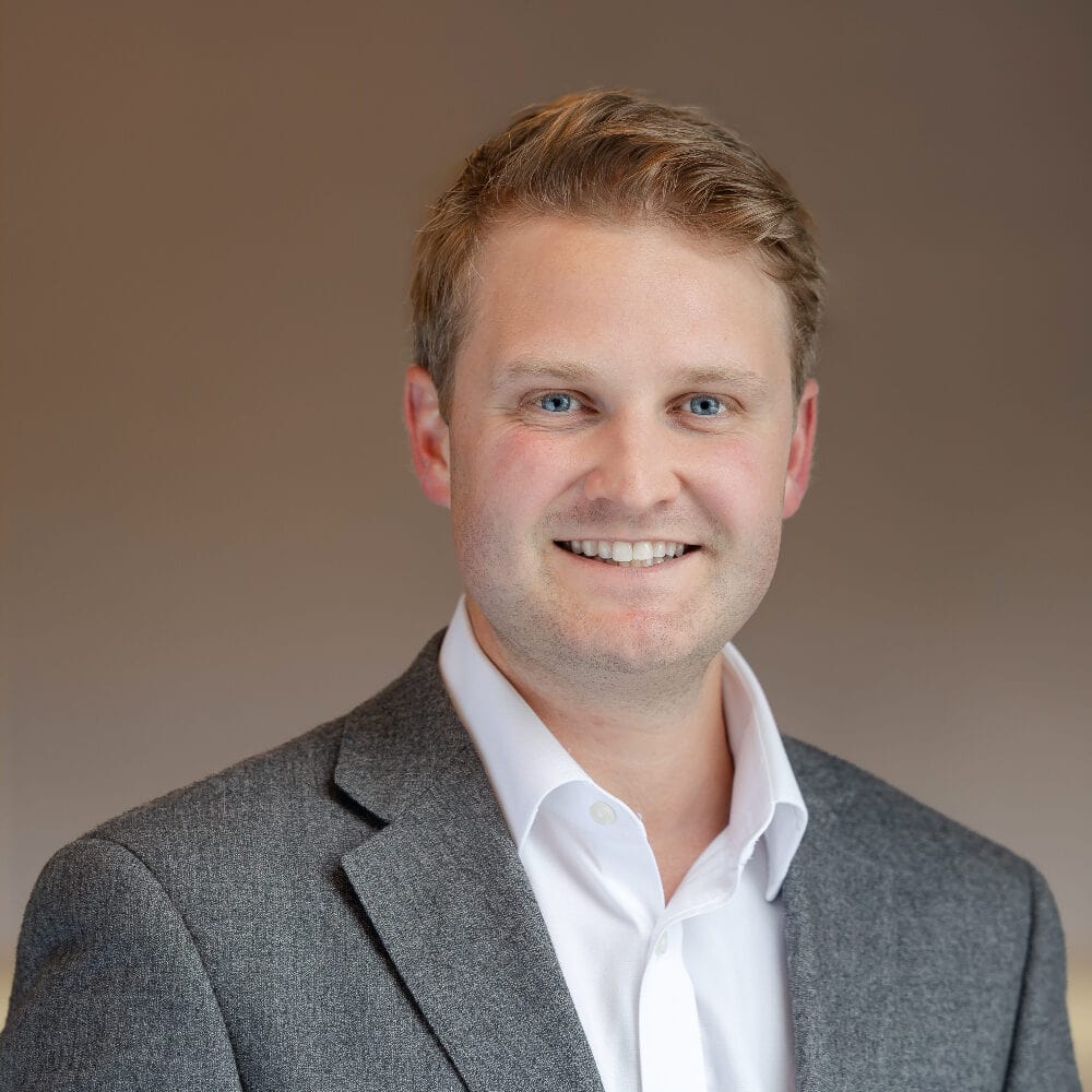 Headshot of a smiling young man with light brown hair and blue eyes, wearing a grey blazer and white shirt.