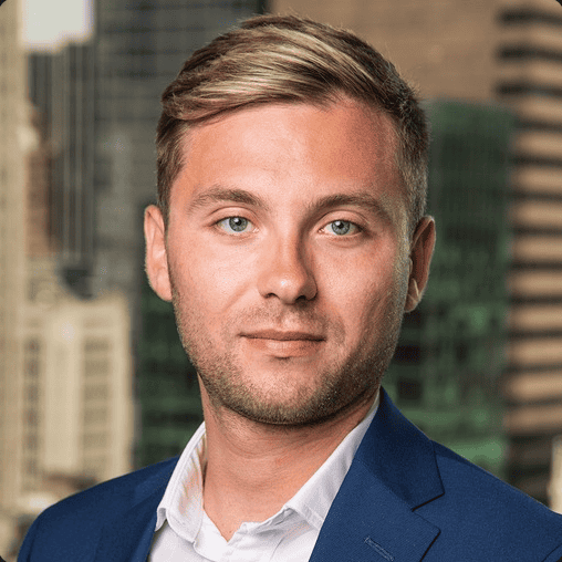 Headshot of a young man with blue eyes and blonde hair, wearing a blue suit jacket and white shirt, against a blurred city background.