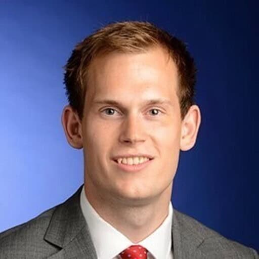 Headshot of a young man with light brown hair, blue eyes, wearing a grey suit and red tie, smiling against a blue background.