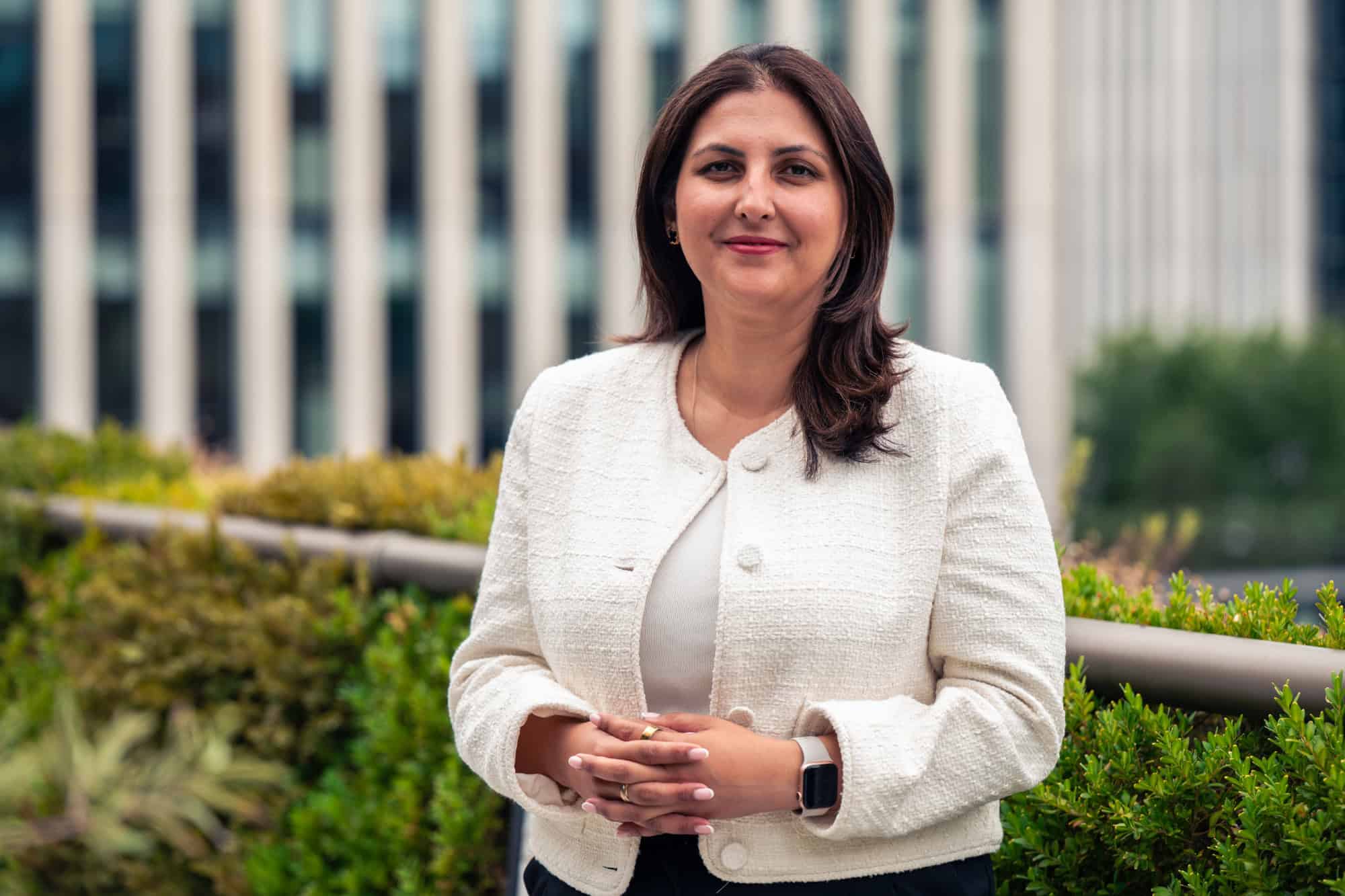 A smiling woman with dark hair and a cream jacket stands outdoors with hands clasped, against a blurred green and building background.
