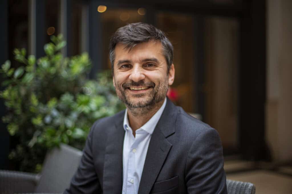 A smiling man with dark hair and a beard wearing a dark suit jacket and light blue shirt.