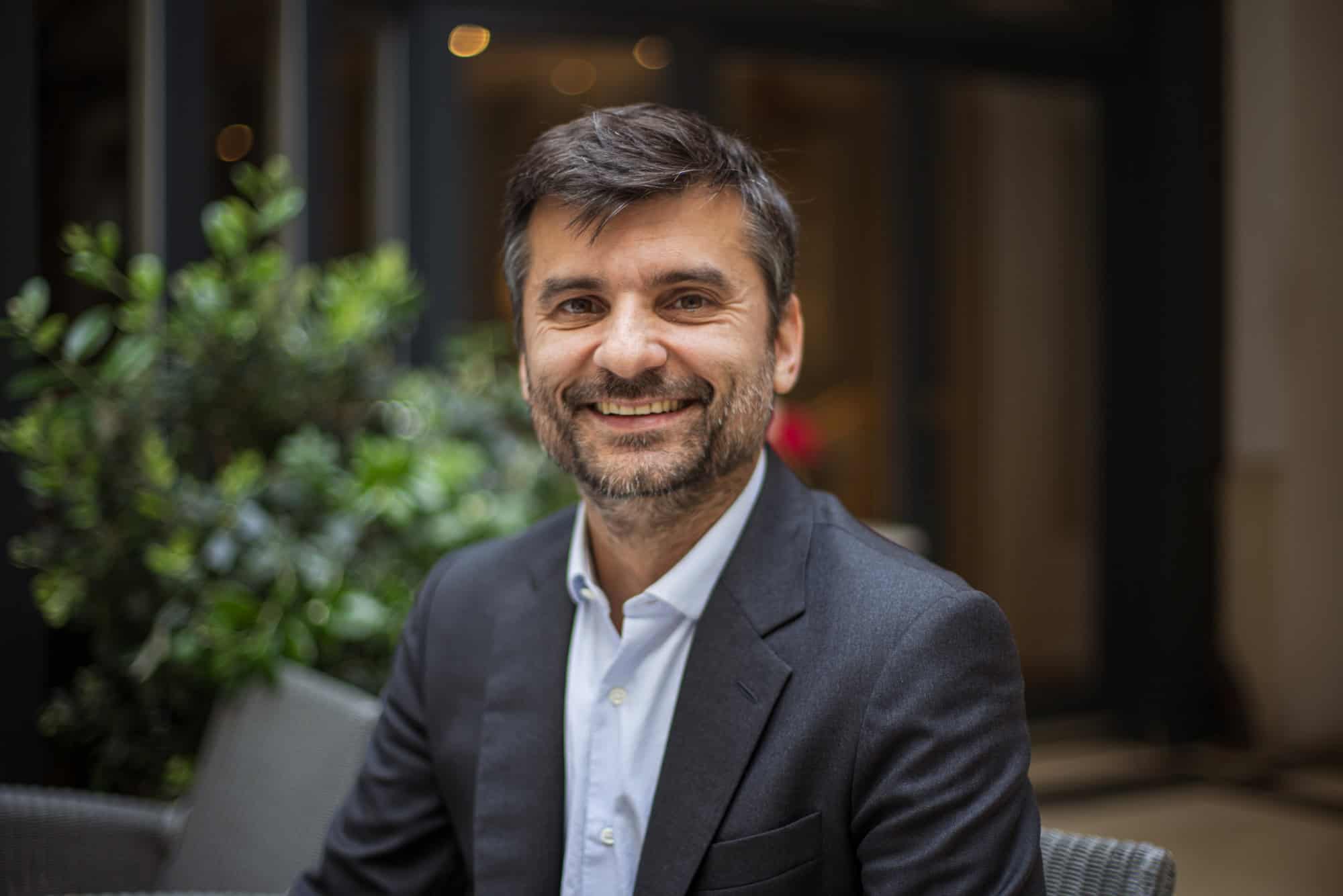 A smiling man with dark hair and a beard wearing a dark suit jacket and light blue shirt.