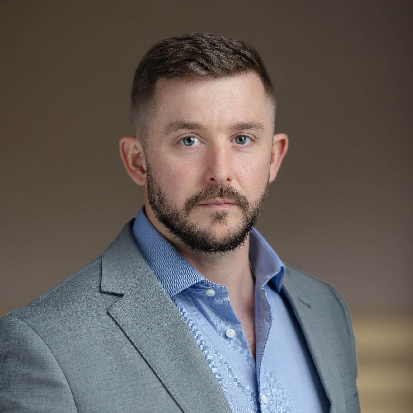 Headshot of a man with blue eyes, short dark hair, and a beard, wearing a grey suit jacket and blue shirt.