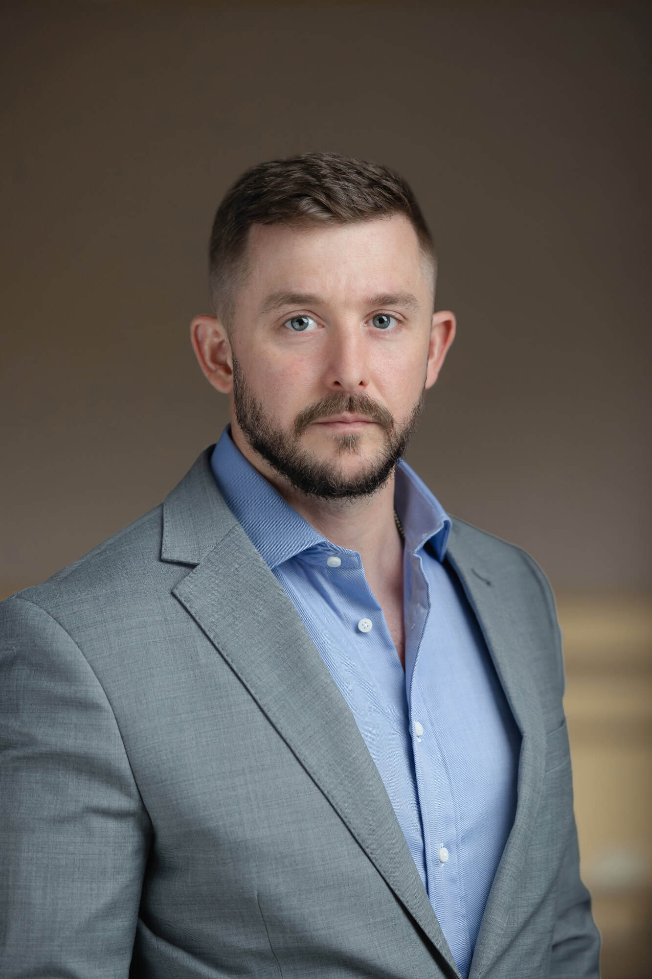 Headshot of a man with blue eyes, short dark hair, and a beard, wearing a grey suit jacket and blue shirt.