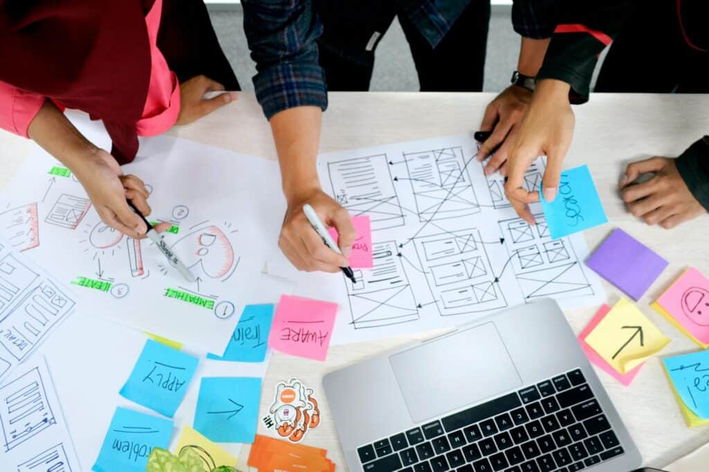 Overhead view of three people's hands collaborating on a table with design documents, wireframes, colorful sticky notes, and an open laptop.