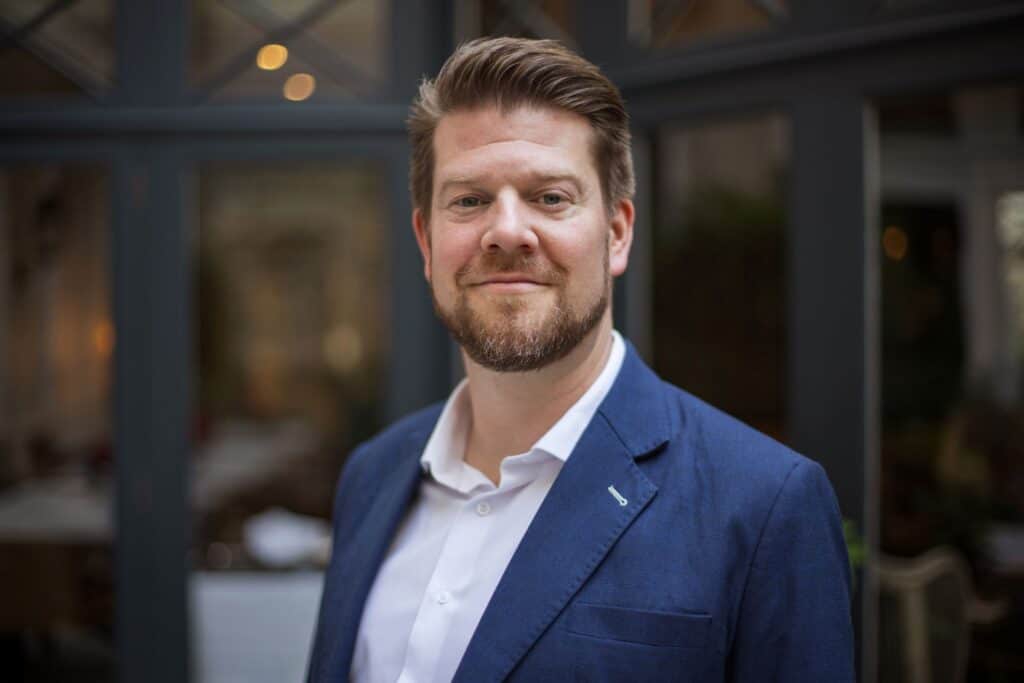 Headshot of a smiling man with light brown hair and beard, wearing a blue blazer and white shirt, against a blurred background.