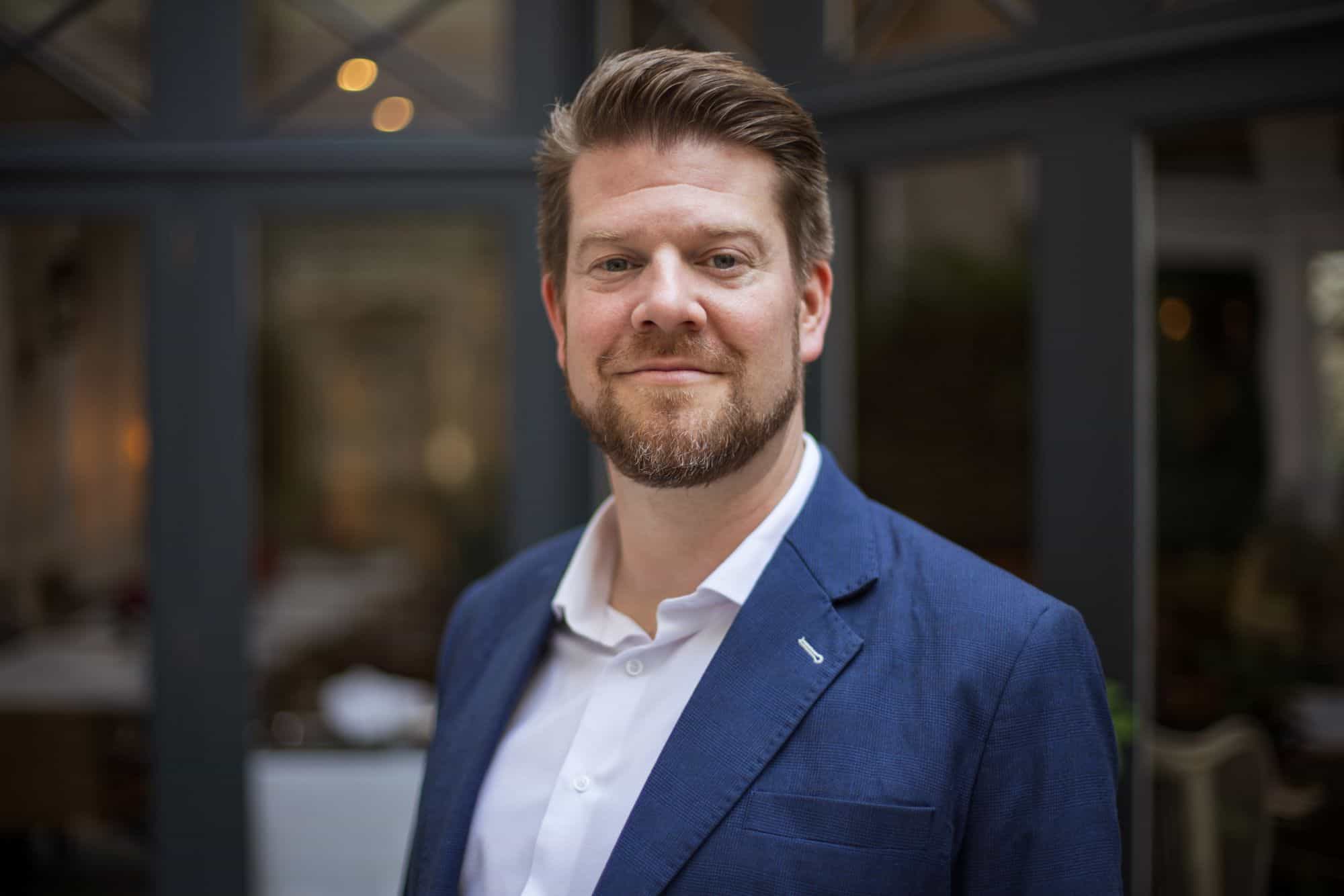 Headshot of a smiling man with light brown hair and beard, wearing a blue blazer and white shirt, against a blurred background.