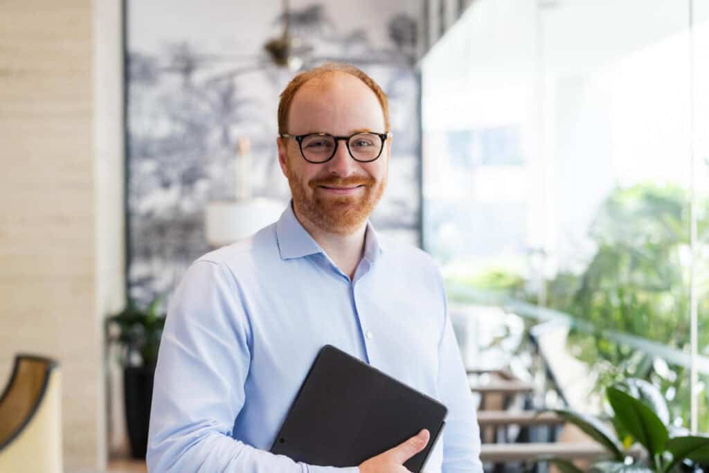 A smiling man with red hair, a red beard, and glasses, wearing a light blue shirt, holds a black tablet in a modern indoor setting.