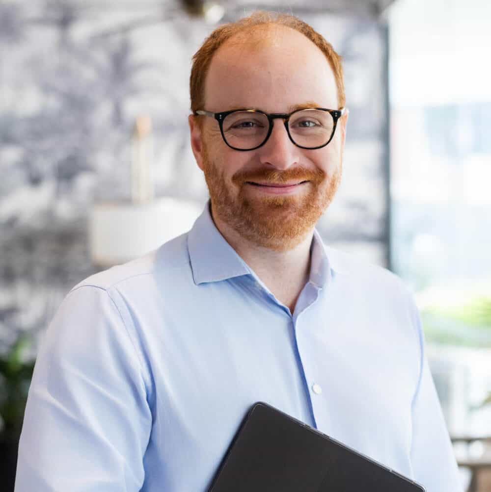 A smiling man with red hair, a red beard, and glasses, wearing a light blue shirt, holds a black tablet in a modern indoor setting.