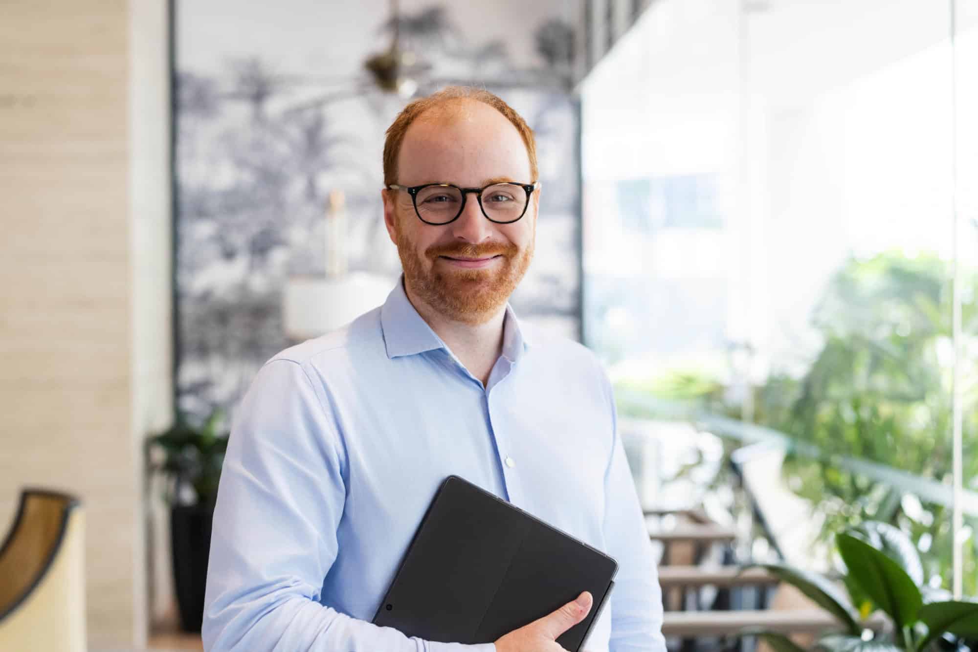 A smiling man with red hair, a red beard, and glasses, wearing a light blue shirt, holds a black tablet in a modern indoor setting.