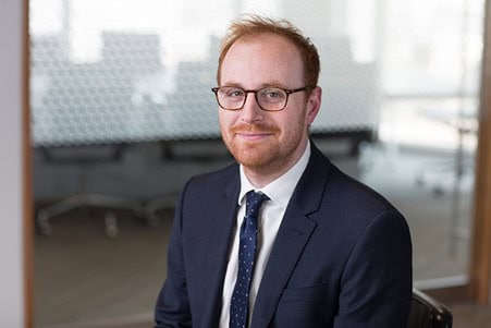 Headshot of a man with red hair and beard, wearing glasses, a dark suit, and a tie, smiling.