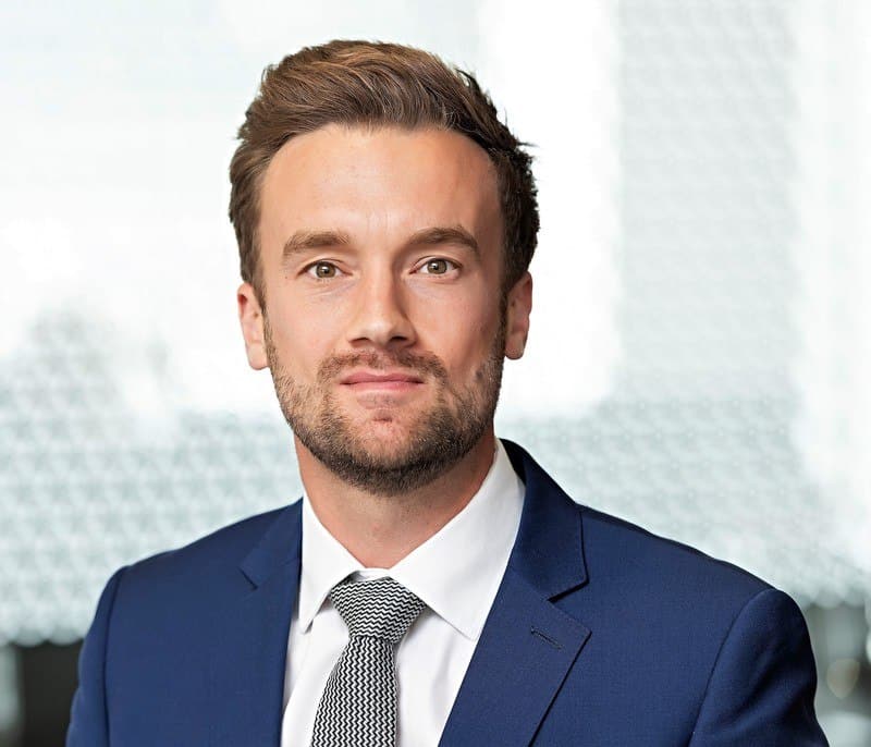 Headshot of a man with light brown hair and beard, wearing a dark blue suit and patterned tie, looking forward.