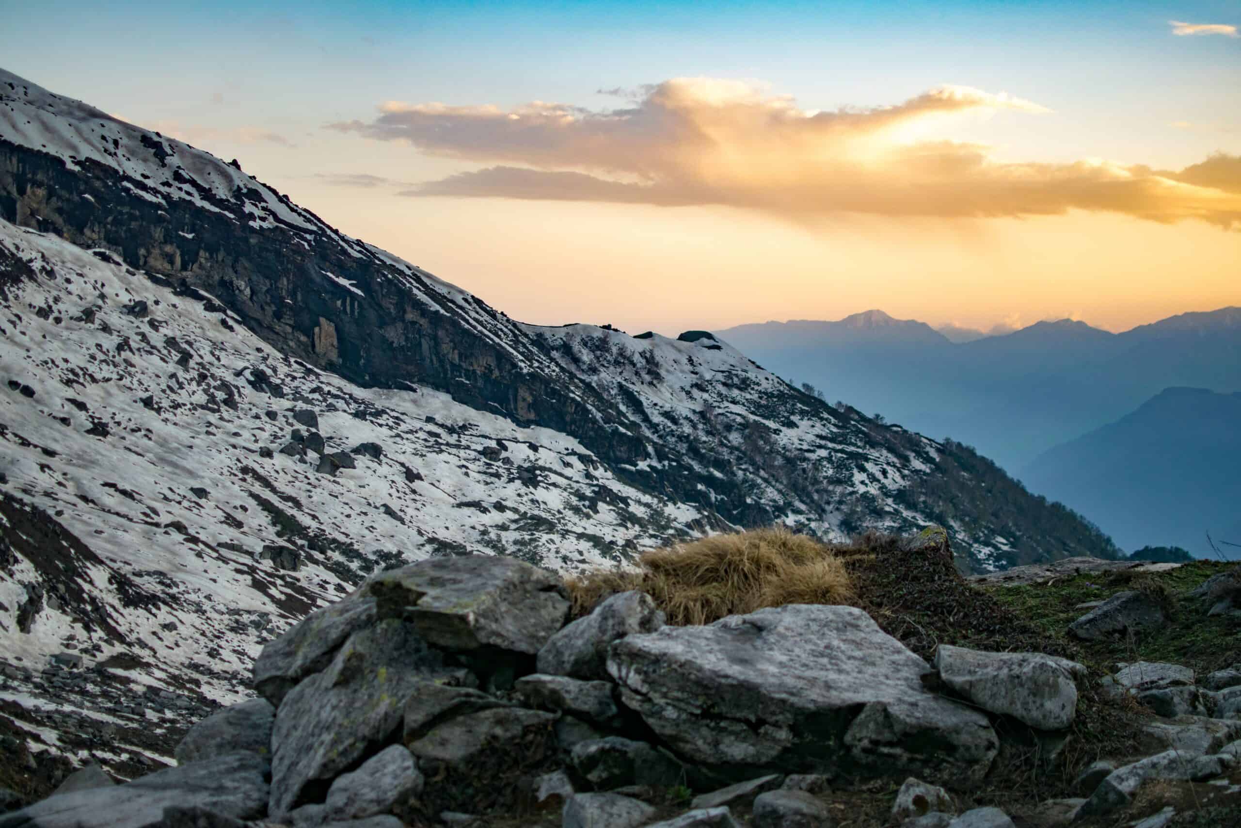 A mountain landscape at sunrise or sunset, featuring snow-covered rocky slopes in the midground, foreground rocks and dry grass, and distant hazy blue mountains under a sky with golden clouds and a blue top.
