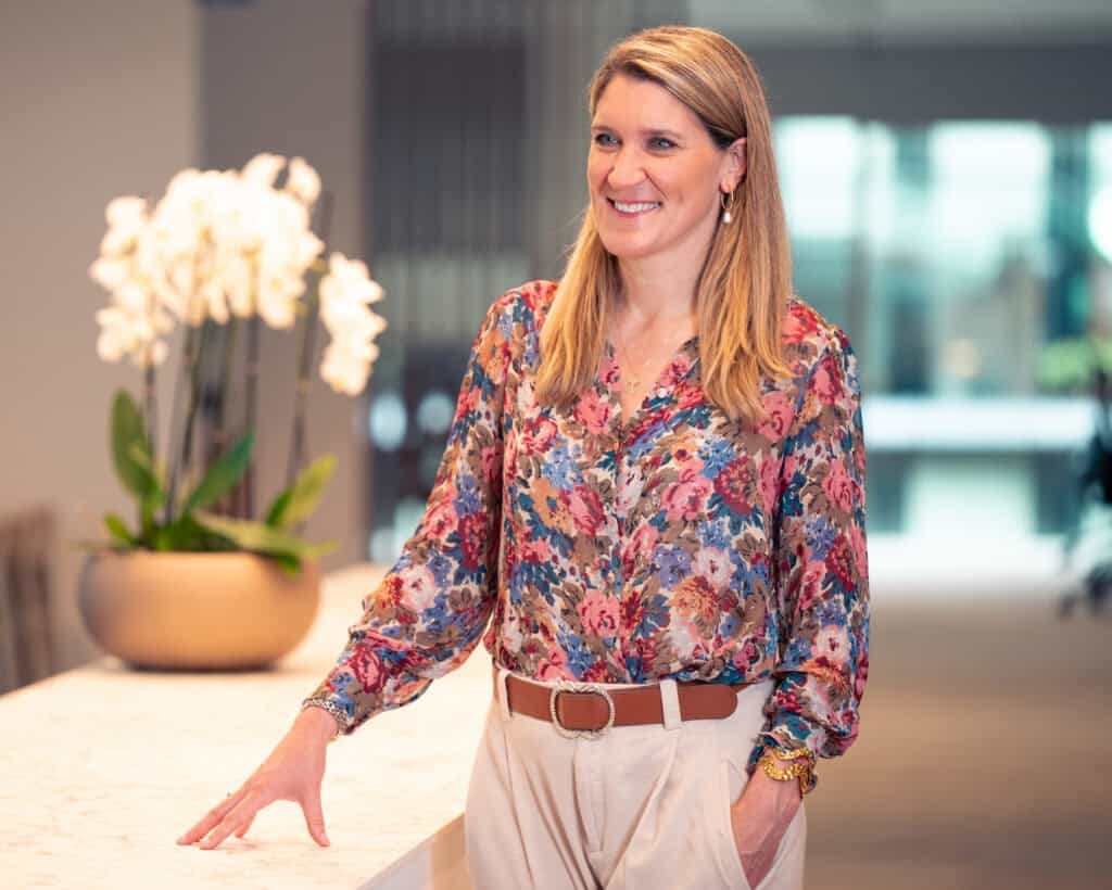 A smiling woman with blonde hair, wearing a floral blouse and light trousers, stands and leans on a table with an orchid plant in the background.