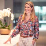 A smiling woman with blonde hair, wearing a floral blouse and light trousers, stands and leans on a table with an orchid plant in the background.
