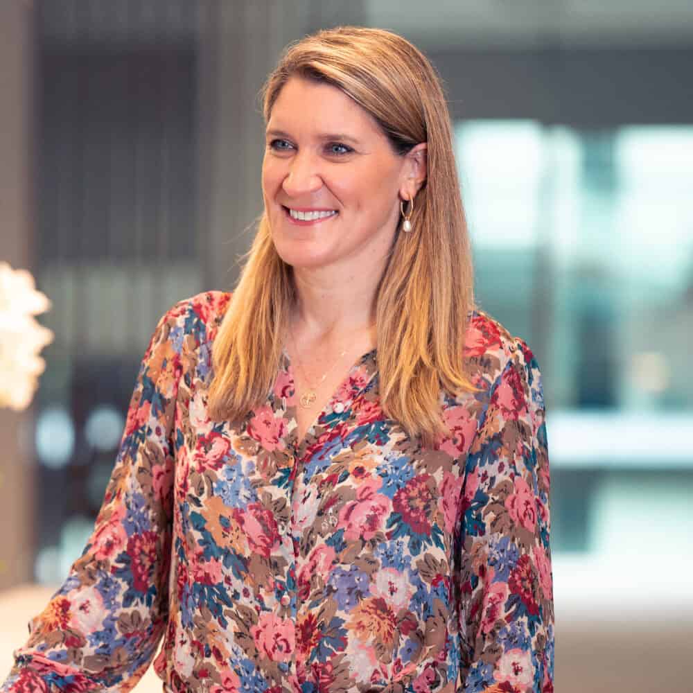 A smiling woman with blonde hair, wearing a floral blouse and light trousers, stands and leans on a table with an orchid plant in the background.