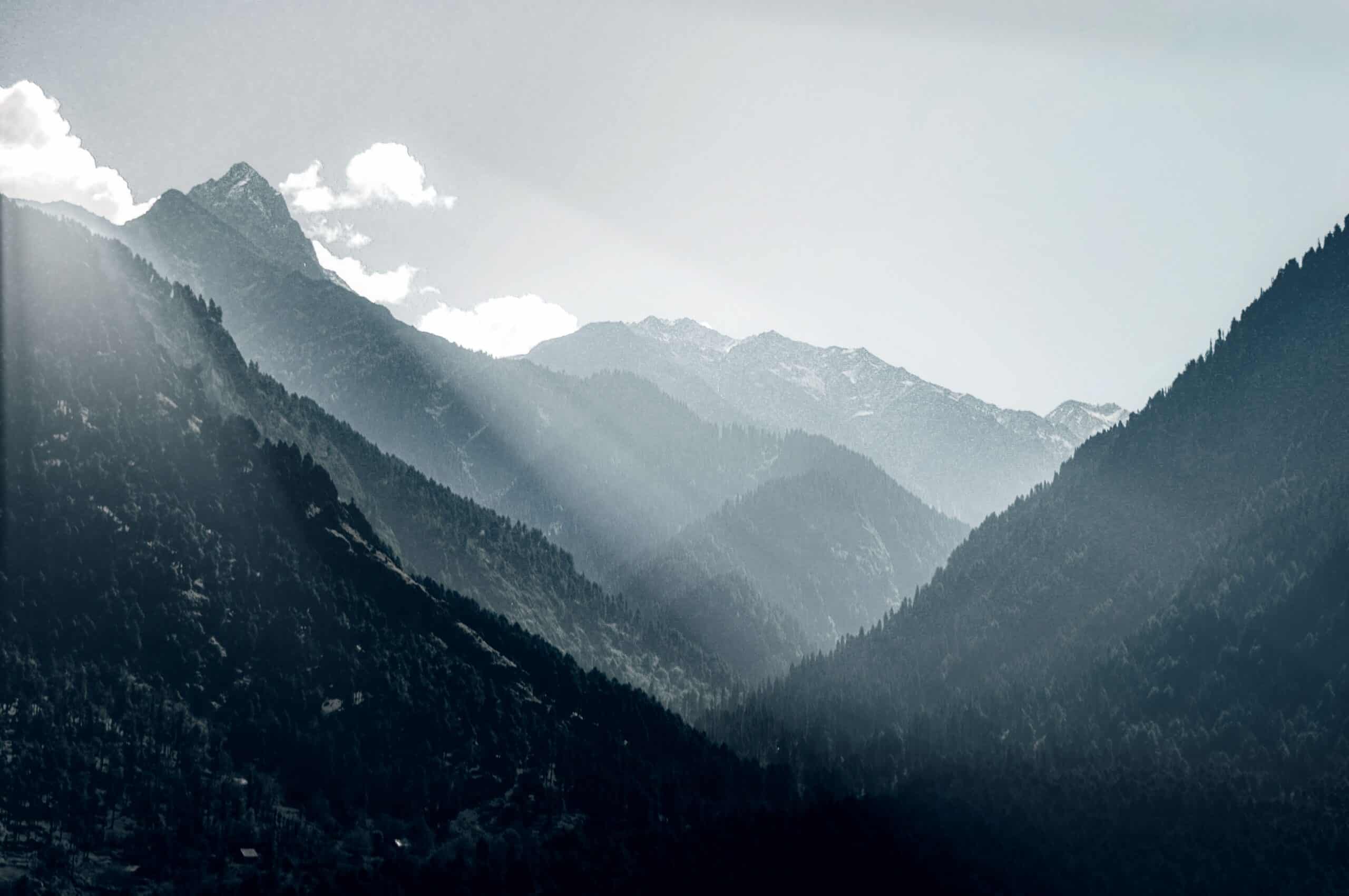 Panoramic view of a layered mountain range covered in evergreen trees, with sun rays breaking through haze.