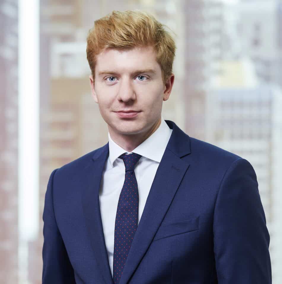 Headshot of a young man with red hair and blue eyes, wearing a dark blue suit and tie, against a blurred city background.